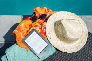 Flat lay of summer essentials by the pool with e-reader, sunhat, and vibrant towel.